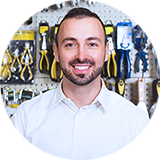 A smiling man dressed in a professional white shirt standing in front of a shelf full of tools.