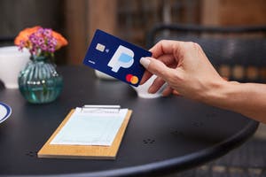 Hand holding a PayPal Cashback Mastercard over a restaurant table with a bill on a clipboard.