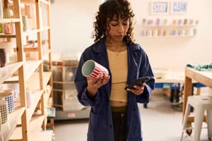 A woman in a pottery studio holds a striped ceramic cup in one hand while checking her phone, surrounded by shelves of handmade ceramics. 