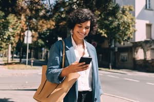 A woman on the sidewalk smiling at her phone as she receives an international money transfer. 