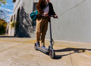 Person in professional clothes riding an electric scooter through the streets with a duffle bag.