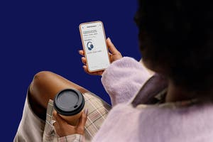 A person sitting and holding a coffee cup looks at a smartphone displaying a shopping app with headphones on the screen against a solid blue background.