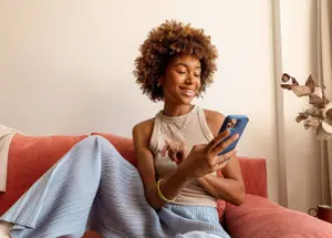 A woman sits on an orange couch using her phone to shop online for holiday gifts