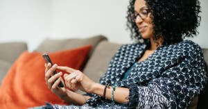 Woman wearing blue sweater sitting on couch and using phone for sales