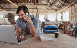 Hombre sonriente en un taller usando su computadora mientras habla por teléfono.