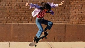 Skateboarder in a purple jacket doing a jump on her skateboard in front of a brick wall.