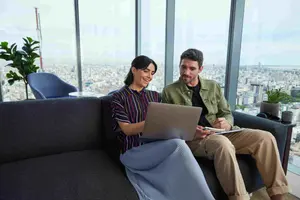 Dos compañeros de trabajo están hablando; la mujer muestra algo en la pantalla de su ordenador mientras el hombre toma notas en un cuaderno.