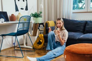 A young woman sitting on the floor, using a smartphone and holding a credit card, in a cozy room with a desk, a chair, and a couch. A large wall art piece is visible in the background.