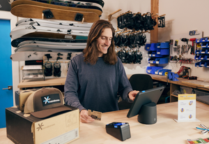 Small business owner checking out a customer purchasing a sweatshirt using the PayPal POS app on an iPad stand with a connected PayPal card reader at a retail counter.