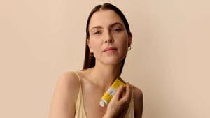 Eine Frau mit zurückgekämmten Haaren hält eine gelbe Creme-Tube an ihre Schulter vor einem beigen Hintergrund.