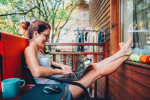 Frau mit Laptop auf dem Schoß sitzt entspannt auf dem Balkon und erledigt eine Online-Überweisung ins Ausland.