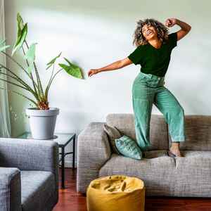 A fashionable woman jumping happily on her modern couch; beside her is a large plant on a glass side table and yellow ottoman