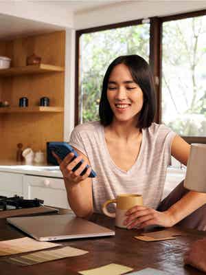 Young millennial woman checking her smartphone with laptop displaying on kitchen countertop.
