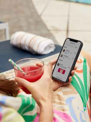 A woman on a lounge chair holding a tropical drink and a mobile phone, mobile phone shows transactions on her PayPal account.