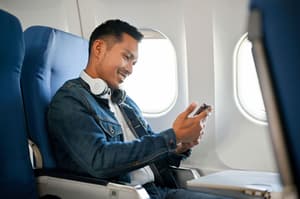 A smiling man sitting in an airplane window seat, wearing a denim jacket and white headphones around his neck, looking at his smartphone.