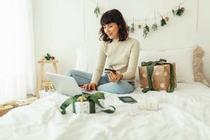 Woman sitting on her bed, surrounded by Christmas trees and holding a credit card, doing online shopping