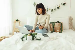 Mujer sentada en su cama, rodeada de decoración navideña y con una tarjeta de crédito en la mano, haciendo compras por Internet.