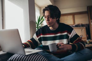 Junger Mann im gestreiften Pullover mit einer Tasse in der Hand, der einen Laptop zu Hause benutzt