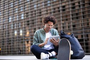 Man sitting outside with a skateboard and looking on his tablet