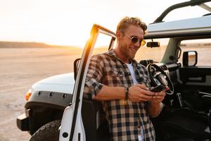 Man standing in the open car door of his Jeep in the sunset, looking at a phone and smiling