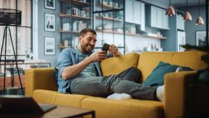 Man in denim shirt lounging on a yellow sofa and smiling at his smartphone