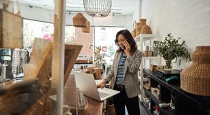 Woman standing behind the counter at a shop, on the phone and looking at an open laptop