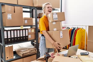 Young blonde woman working at small eCommerce business, holding a pile of boxes for shipping
