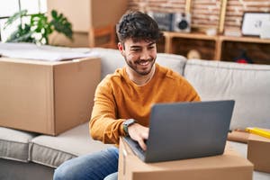 Young smiling hispanic man sitting on couch using laptop at new home