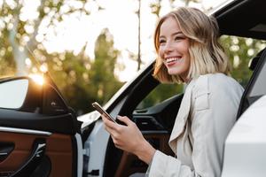 Smiling young woman holding her phone and getting out of the driver's seat of a grey SUV.