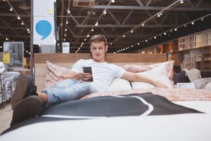 Young man lying on a bed at a furniture store while looking at his phone.