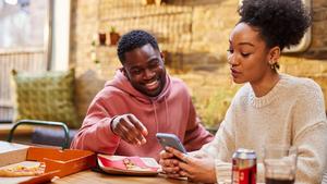 A man and a woman look at a smartphone while sitting at a table, eating pizza