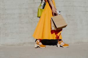 A person in an orange dress and tan platform sandals walking while carrying brown paper shopping bags and a smartphone, illustrating the blend of digital and in-person retail.