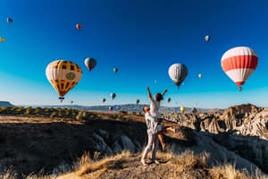 A man lifts a woman with raised arms in a joyful, celebratory embrace on a rocky hilltop with dozens of colorful hot air balloons floating across a vivid blue sky.