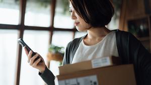 A woman holds a package as she looks at her smartphone. 
