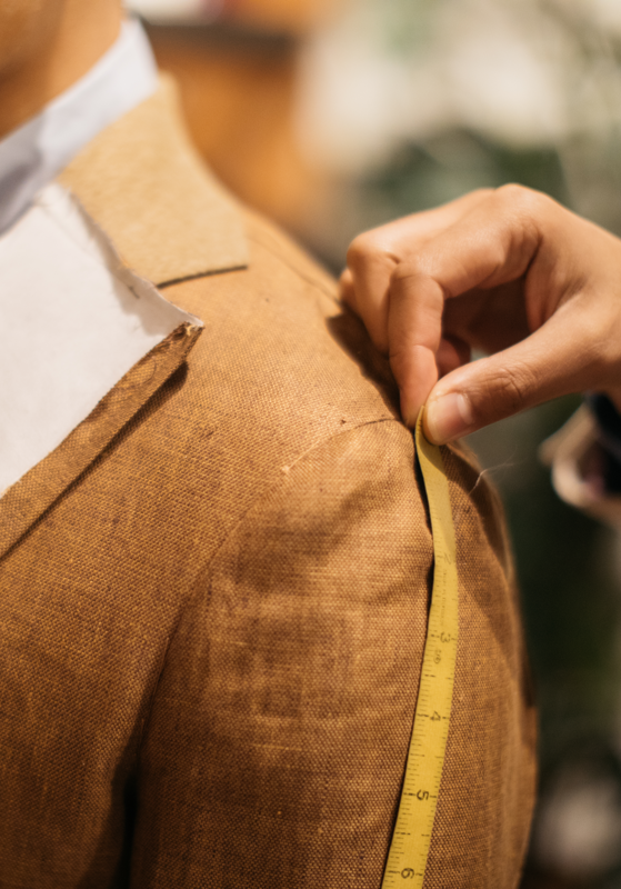Closeup of a tailor’s hand measuring the shoulder of a customer wearing an unfinished jacket prototype.