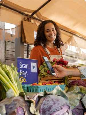 Eine lächelnde Frau an ihrem Marktstand mit einem PayPal-QR-Code-Schild, eine einkaufende Person scannt den QR-Code mit ihrem Handy