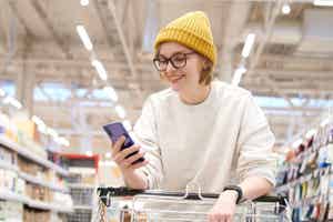 A smiling woman looking at her smartphone while holding onto a grocery cart.