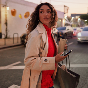 Woman wearing long beige coat and holding a phone, walking through traffic.
