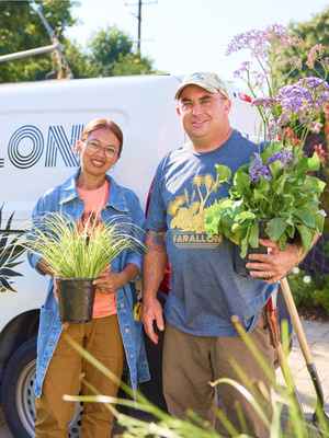 2 happy landscaping business owners; 1 is holding an ornamental grass, 1 is holding a large flowering plant