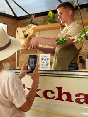 A woman wearing a hat scans a QR code at a mobile plant store; the merchant packages the plant she just purchased