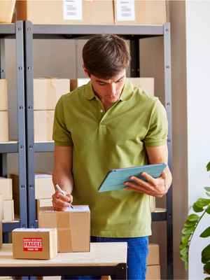 Man in a sage green shirt and jeans looks at his tablet while writing out a shipping label on a small box
