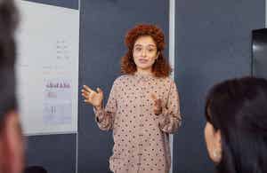 Person with curly hair giving a presentation, gesturing with hands. A whiteboard with charts and notes is in the background.