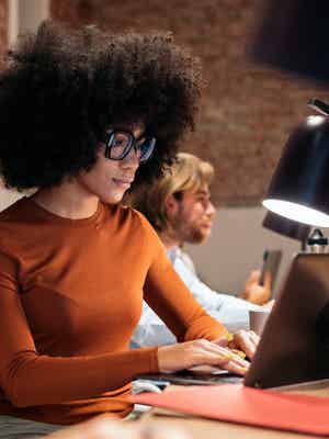 Person working on a laptop at a shared table style desk, with another person at their workspace behind them in a converted warehouse style office.