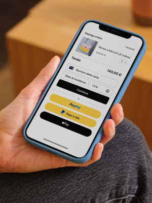 Persona che tiene un telefono mostrando schermata di checkout con opzioni di pagamento inclusi carta di debito/credito, PayPal, Paga a rate e Apple Pay