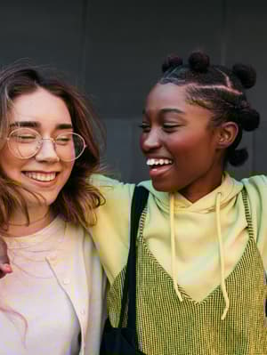 Three people smiling together, with the person in the middle placing their arms around the shoulders of the other two. All are dressed in casual clothing.