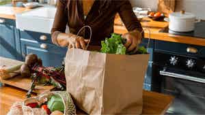 Una persona in cucina svuota una borsa di carta della spesa piena di verdure a foglia verde; sul tavolo di legno ci sono delle verdure e una borsa a rete con altri prodotti freschi, con una cucina moderna sullo sfondo.