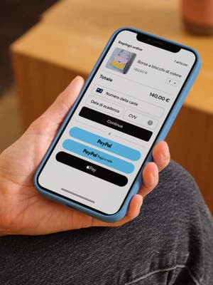 Persona che tiene un telefono mostrando schermata di checkout con opzioni di pagamento inclusi carta di debito/credito, PayPal, Paga a rate e Apple Pay