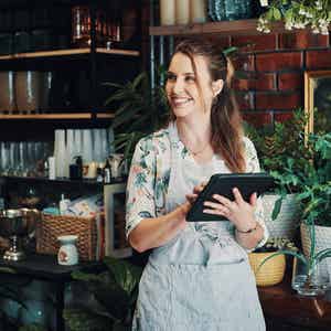Smiling small business owner in a floral top and linen apron holds a tablet while standing in her plant and homeware shop, surrounded by lush potted greenery, glass vases, and a warm exposed brick wall backdrop