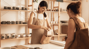 Pottery shop owner packing a handmade ceramic product into a paper bag for a customer.