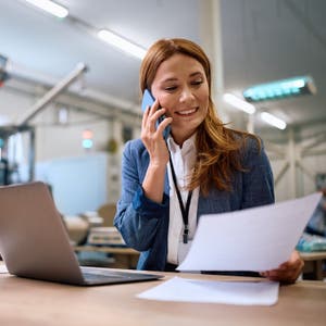 Businesswoman in a manufacturing facility talking on her phone while reviewing a printed document next to an open laptop.